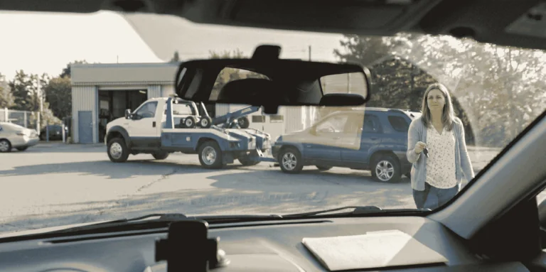 Here is a short, descriptive alt text option for the image **Obvi-blog3-June.jpg**: > View from inside a car dashboard looking out at a woman walking forward while holding keys, with a tow truck preparing to hook up a blue SUV outside an auto repair shop in the background.